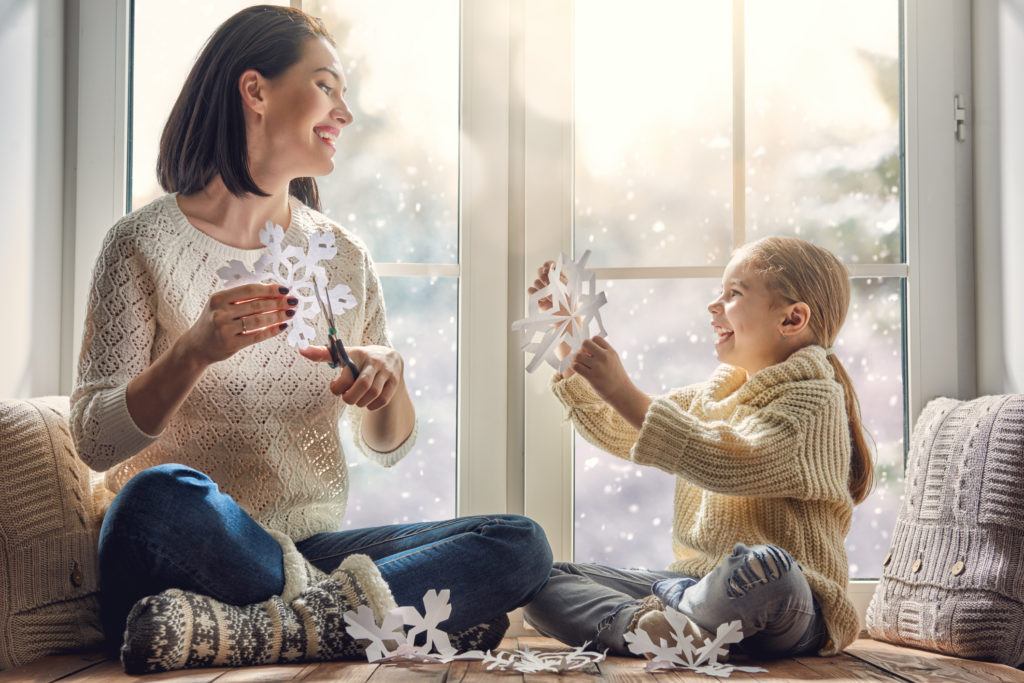 mother and daughter sit by a snowy window happily making paper snow flakes